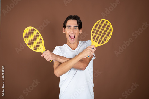 Funny guy with children's tennis rackets posing on a brown background. © vladorlov