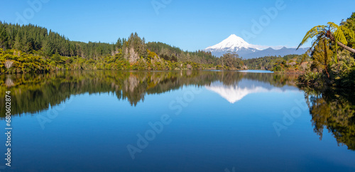 The snow-covered summit of Mt Taranaki as well as lush native trees and bushes reflected in the calm waters of Lake Mangamahoe on New Zealand's North Island