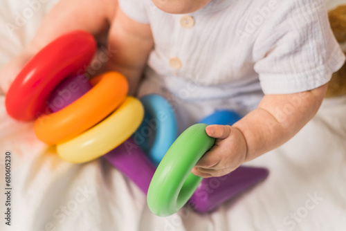 Close-up baby playing with colorful rainbow toy pyramid on white sunny bedroom. Toys for little kids. Child with educational toy.