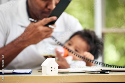 Young Asian father holding calling telephone and treat baby boy feeding milk and working from home office as blur background photo