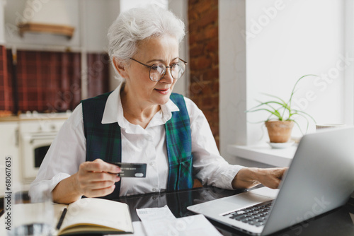 Happy smiling senior female with gray hair in formal wear sitting at kitchen table holding plastic card in hands doing online payment using laptop and banking application, paying utility bill photo