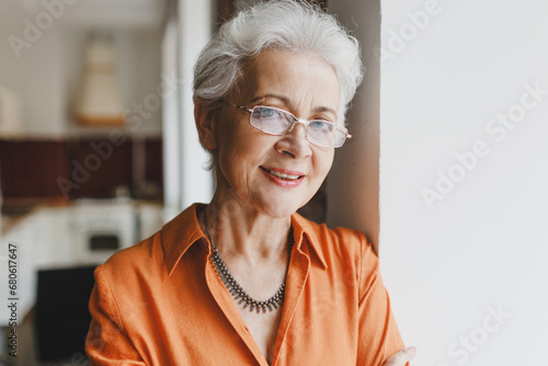 Closeup indoor portrait of elderly wise kind female grandmother in glasses with gray hair in orange shirt leaning against wall at home on blurred kitchen background, looking at camera with smile