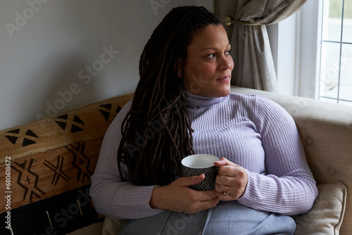 Biracial pregnant woman with locs looking out of window and holding tea cup. Concept of family, pregnancy, couple, diverse, motherhood