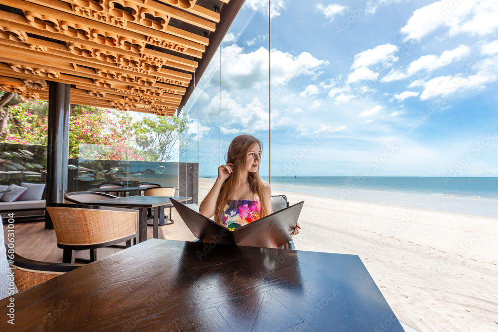 Relaxed female tourist enjoying sea breeze while looking at menu, beachfront dining establishment, vacation leisure.