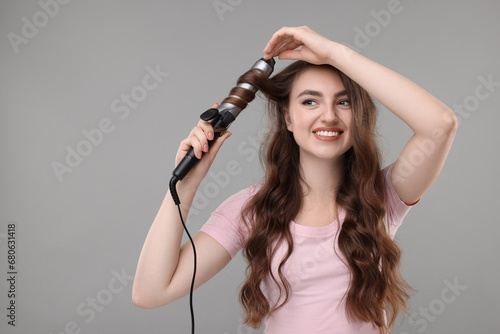 Beautiful young woman using curling hair iron on grey background, space for text