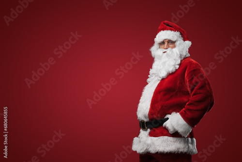Jolly Santa Claus in traditional red suit standing with hands on hips against red backdrop