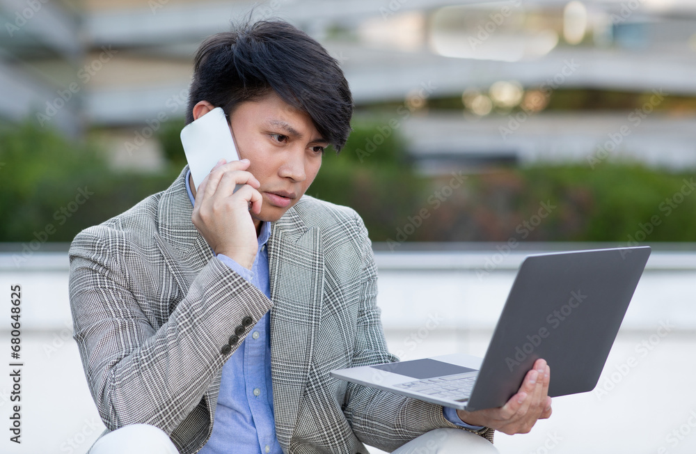Displeased Korean manager man using laptop talking with phone outdoors Displeased Korean manager man using laptop talking with phone outdoors