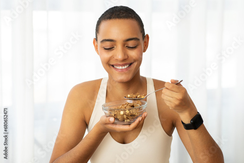 Smiling millennial latin lady in sportswear eat muesli cereal, enjoy breakfast