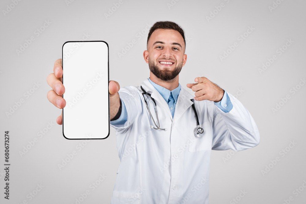 Smiling male doctor pointing at phone with blank screen Smiling male doctor pointing at phone with blank screen