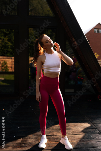 Portrait of athletic female wearing sportwear rotating neck warming up before morning exercise, muscles warm up to prevent injury from workout, standing on background of house on sunny summer morning.