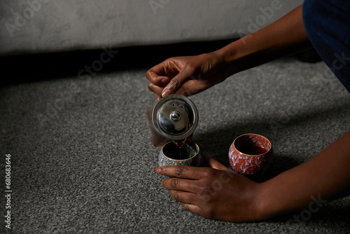 Young black woman pouring tea into unique handcrafted cups on the floor of living room