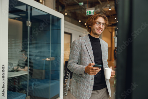 Smiling businessman use mobile phone and drink coffee while standing on coworking background