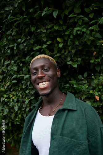 Stylish man with blonde hair smiles to camera outdoors in front of greenery