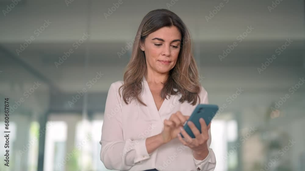 Smiling mid aged business woman executive using smartphone in office. Happy elegant mature female employee or professional bank sales manager looking at cell mobile phone tech standing in lobby. Smiling mid aged business woman executive using smartphone in office. Happy elegant mature female employee or professional bank sales manager looking at cell mobile phone tech standing in lobby.