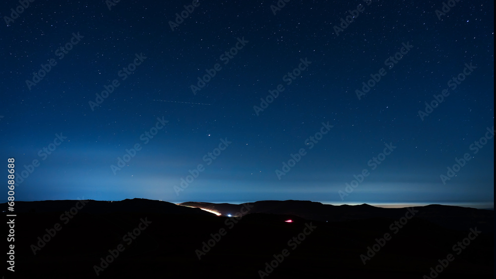 Night starry sky in the mountains. A luminous vehicle is driving along a mountain road. A strip of satellites in the sky among the stars.