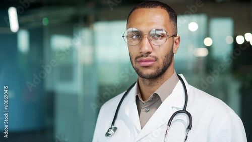 Portrait young serious confident focused doctor looking at camera while standing in modern office clinic close up. Handsome mixed race physician in glasses and white coat posing Head shot doc Closeup photo