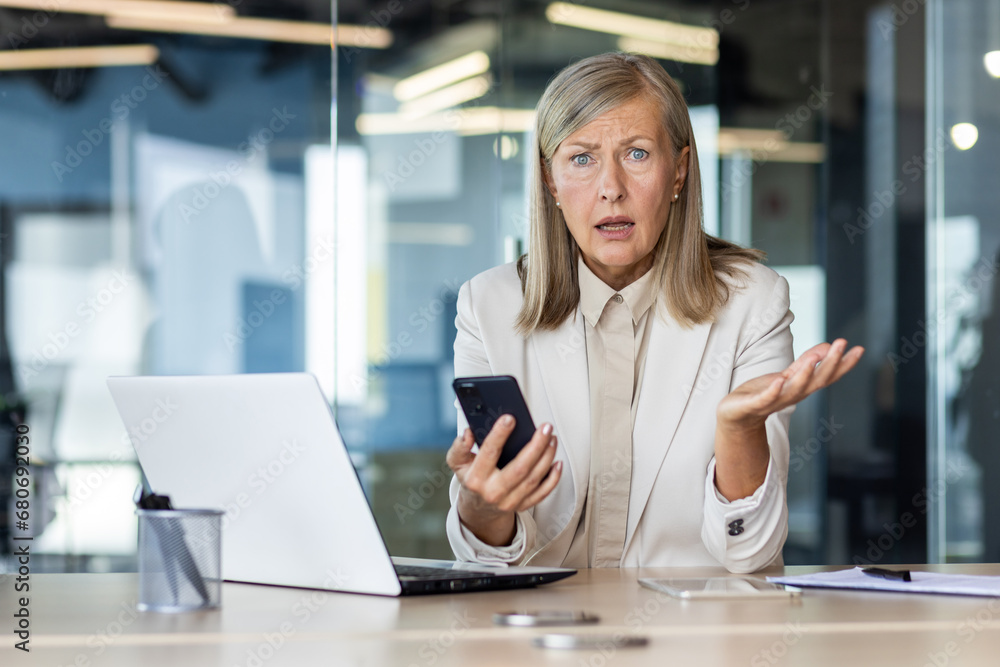Portrait of a worried senior business woman sitting in the office at a laptop, holding a phone in her hand, looking at the camera and throwing her hands in frustration. Portrait of a worried senior business woman sitting in the office at a laptop, holding a phone in her hand, looking at the camera and throwing her hands in frustration.