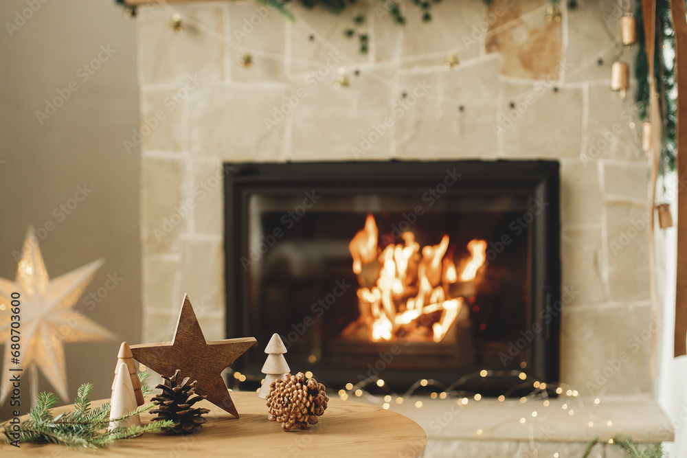Merry Christmas and happy holidays! Stylish christmas wooden trees, star, pine cones and fir branches on table against burning fireplace. Modern rustic eco friendly decor in living room - obrazy, fototapety, plakaty 