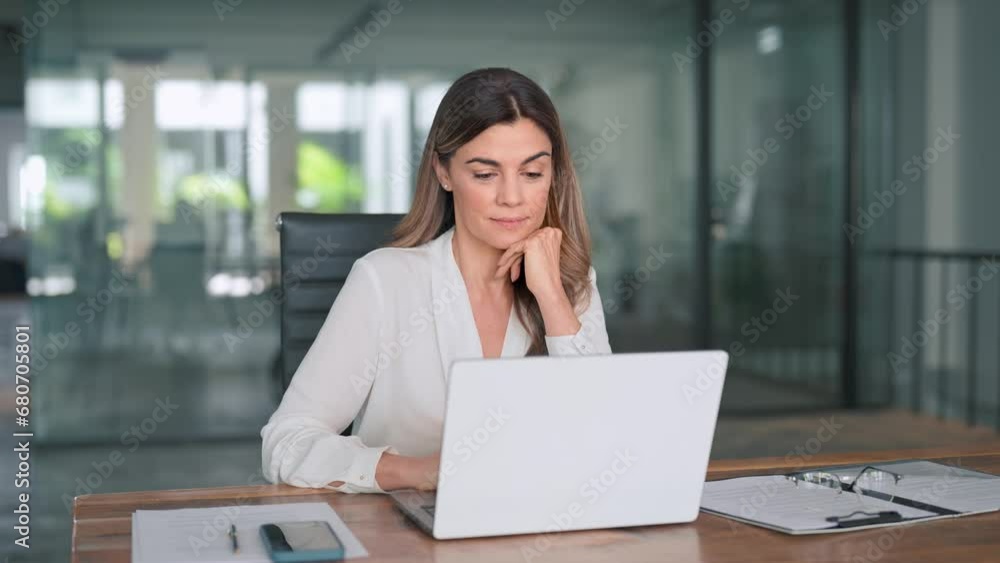 Smiling busy mature middle aged professional latin business woman manager executive looking at laptop computer technology in office working on digital project sitting at desk.