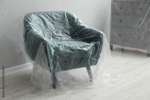 Stylish armchair and chest of drawers covered with plastic film near light grey wall indoors