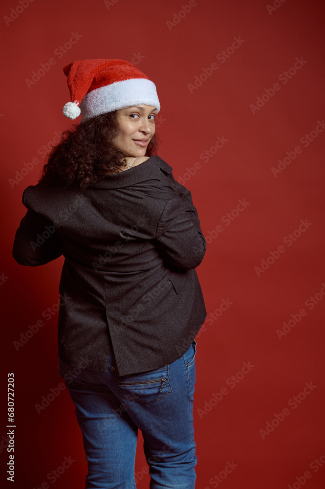 Back view beautiful ethnic young adult pregnant woman wearing Santa hat, smiles cutely looking at camera, red background