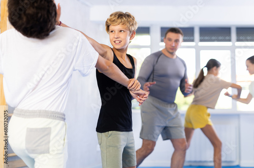 Two preteen boys learn to do power grabs in pairs during a self-defense lesson under the guidance of a trainer in the gym