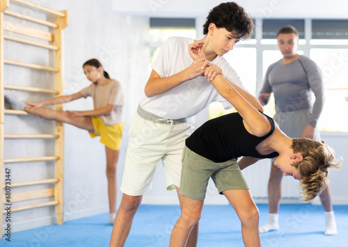 Two preteen boys learn to do power grabs in pairs during a self-defense lesson under the guidance of a trainer in the gym