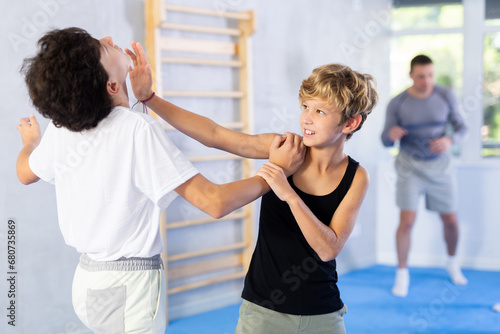 Two preteen boys learn to do power grabs in pairs during a self-defense lesson under the guidance of a trainer in the gym