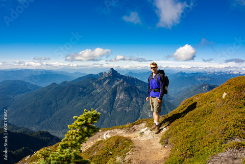 Adventurous athletic male hiker standing on a hiking trail on top of a rugged mountain at at the camera smiling, in the Pacific Northwest with jagged mountains in the background. © Pelo Blanco Photo