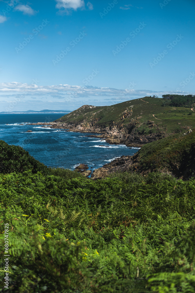 Captivating Ons Island scene: lush greenery in the foreground, azure sea, rocky cliffs, Pontevedra estuary, and a summer sky with scattered clouds from the Buraco do Inferno path.