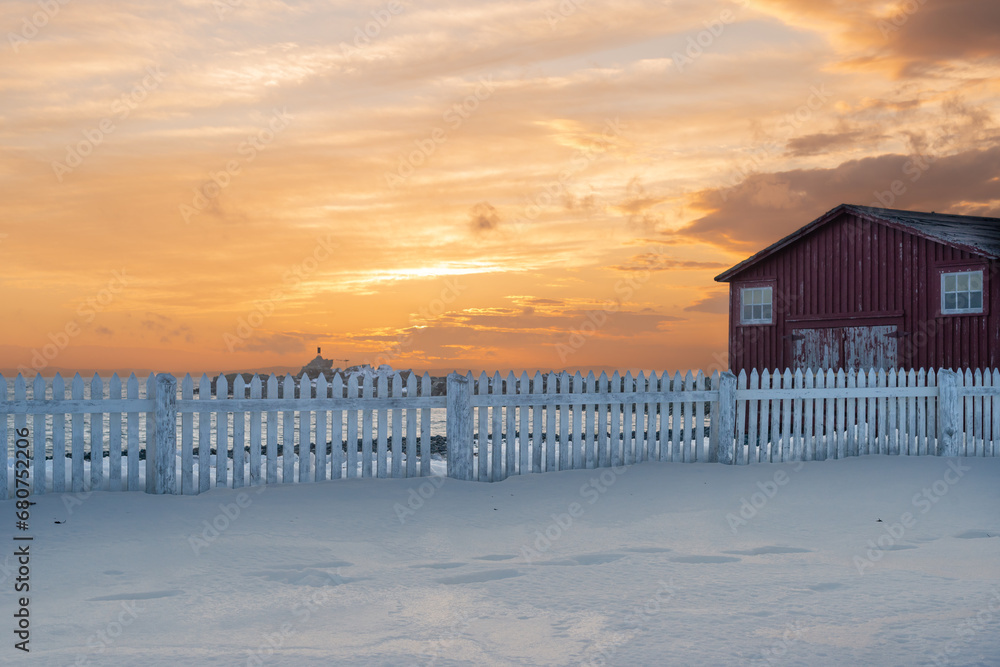 A long white wooden picket fence divides two snow-covered fields on a farm. An old red barn stands in the meadow with a vibrant orange sky and clouds in the background. The sunrise is colorful. A long white wooden picket fence divides two snow-covered fields on a farm. An old red barn stands in the meadow with a vibrant orange sky and clouds in the background. The sunrise is colorful.