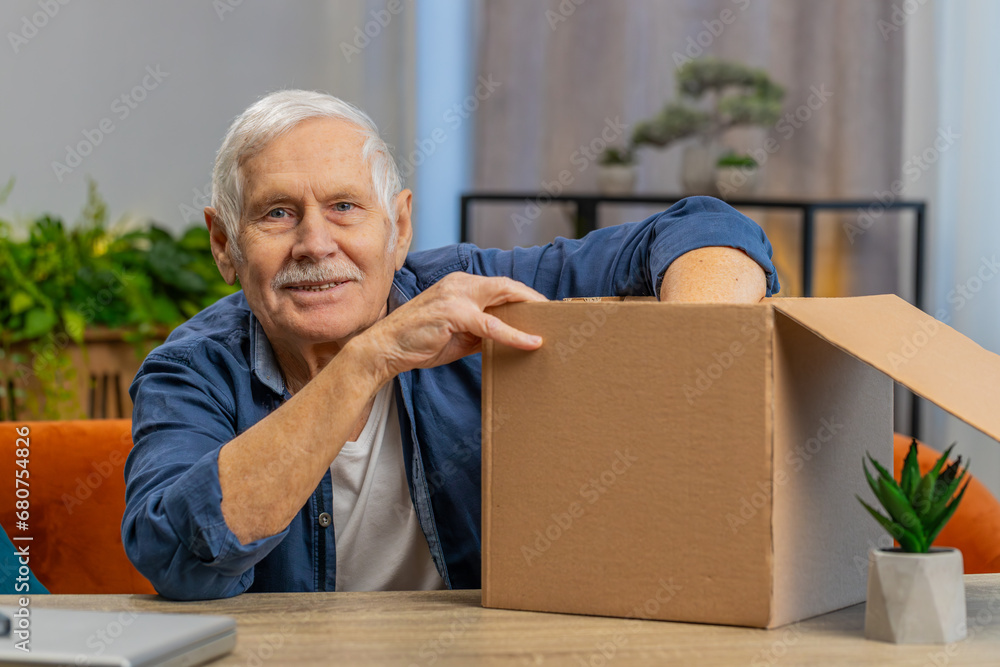 Happy senior mature old man unpacking delivery parcel at home. Smiling satisfied elderly grandfather shopper, online shop customer opening cardboard box receiving purchase gift by fast postal shipping Happy senior mature old man unpacking delivery parcel at home. Smiling satisfied elderly grandfather shopper, online shop customer opening cardboard box receiving purchase gift by fast postal shipping
