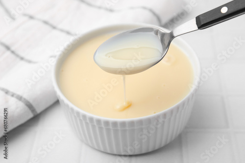 Condensed milk flowing down from spoon into bowl on white tiled table, closeup