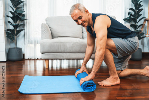 Active and sporty senior man preparing, rolling fitness exercising mat on living room floor at home. Home exercise as concept of healthy fit body lifestyle after retirement. Clout