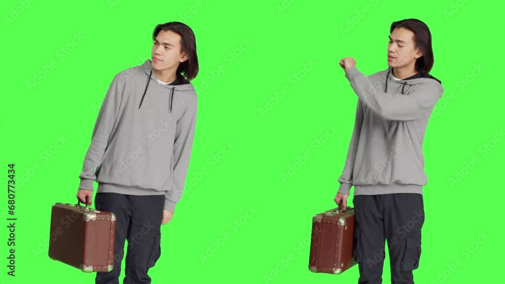 Male model waiting in studio, looking around and checking time on wristwatch. Person carrying vintage suitcase standing over greenscreen backdrop, acting impatient on camera. Male model waiting in studio, looking around and checking time on wristwatch. Person carrying vintage suitcase standing over greenscreen backdrop, acting impatient on camera.