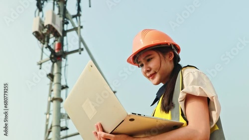 Asian female engineer wearing helmet performs field work telecommunications tower controlling mobile power installation using laptop maintain 5G network installed high rise building.