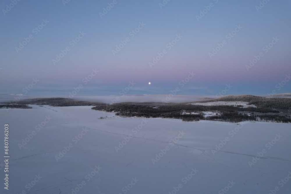 Moon rises over a tranquil, snowy expanse in the fading Arctic light.