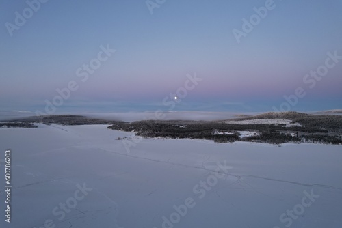 Moon rises over a tranquil, snowy expanse in the fading Arctic light.