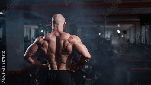 Muscular bald man posing shirtless. Bodybuilder demonstrating back muscles in the gym.