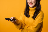 A happy young woman in her 30s, wearing a yellow sweater, sits comfortably on her sofa, watching TV with a remote control.
