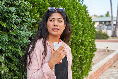 Woman drinks fast food soda with straw in front of hedge inquisitive expression