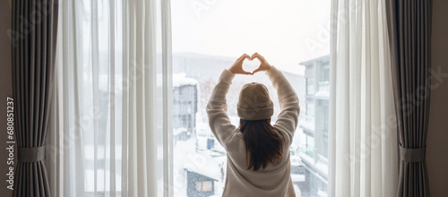 young woman in sweater looking through the window in winter season, happy female rising arms and stretching after waking at apartment or home in the morning. Lifestyle and Relaxing concepts