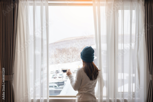 young woman in sweater with cup of coffee looking through the window in winter season, happy female enjoying snowfall outdoor view at apartment or home in the morning. Waking and Relaxing concept