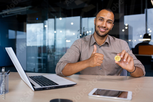 Happy african american businessman sitting in office holding virtual cryptocurrency bitcoin showing like thumb up.