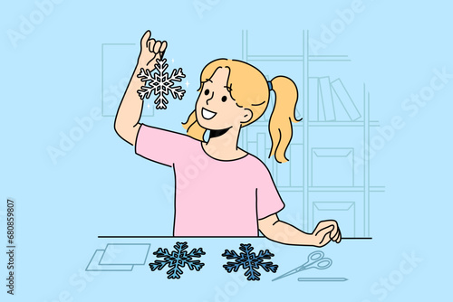 Little girl cuts out paper snowflakes preparing handmade christmas decorations for home on eve of new year. Child stands near table with scissors demonstrating snowflake made with own hands