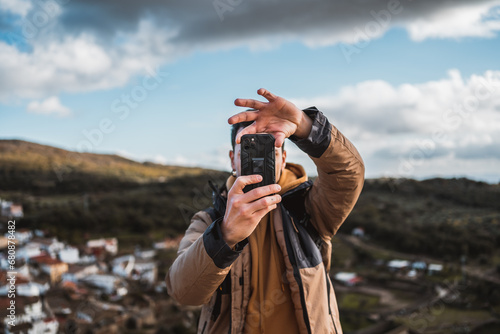 Young happy photographer man traveling sightseeing in a small town, taking a photo with his cell phone of the panoramic view of the town and the mountains, from a castle on a sunny day