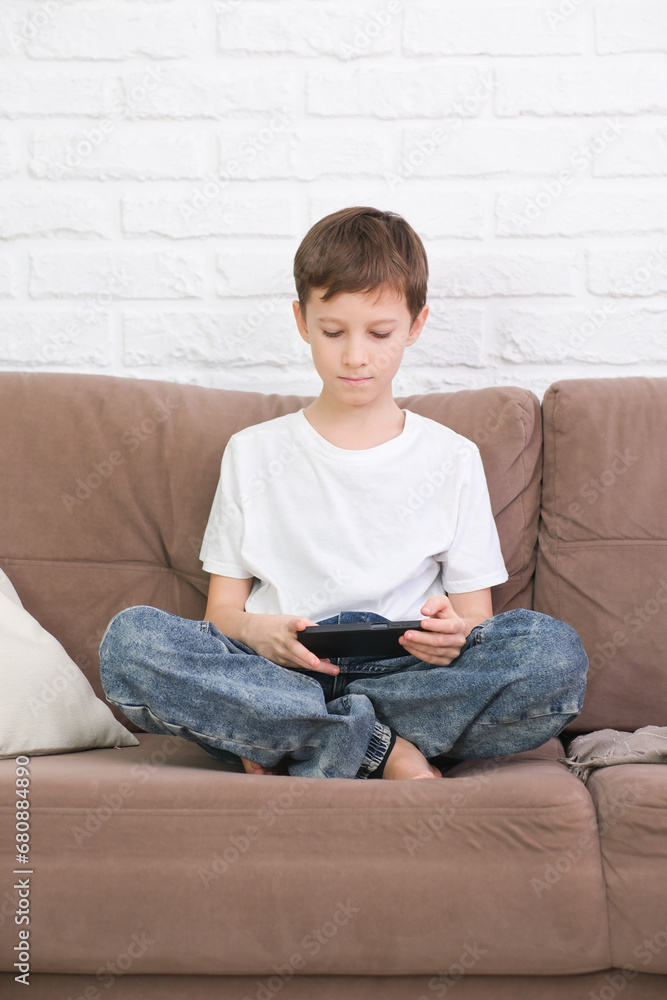 Close up cute little boy wearing headphone using smartphone, looking at screen, child holding phone in hands, sitting on couch at home alone, playing mobile device game, watching cartoons online. Close up cute little boy wearing headphone using smartphone, looking at screen, child holding phone in hands, sitting on couch at home alone, playing mobile device game, watching cartoons online.