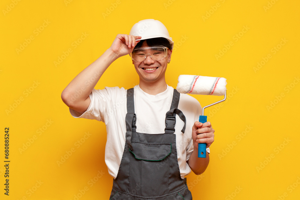 Asian male painter in helmet and overalls holds roller and smiles on yellow isolated background, Korean guy builder