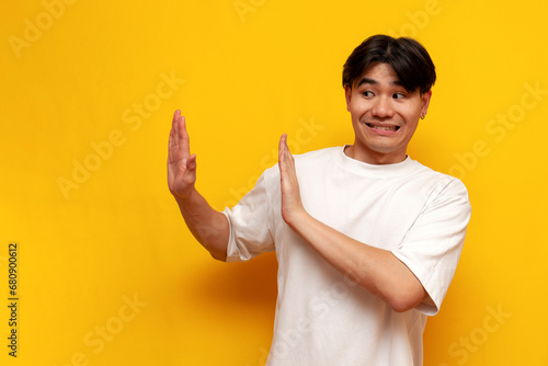 young asian guy in white t-shirt refuses and avoids on yellow isolated background, korean man refuses photo