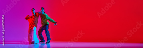 Young man and woman in notion, dancing hip hop against pink red background in neon light. Banner. Concept of hobby, action, street style, contemporary dance, youth, fashion